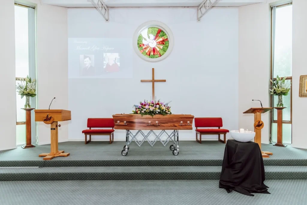 Front sanctuary stage featuring a timber casket with floral spray, wooden lectern, central cross, stained glass dove window, and projected photo tribute screen inside the East Geelong Uniting Chapel at 199 Ormond Rd.