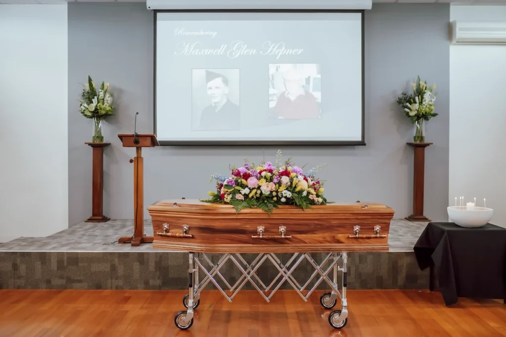 Direct front view of the raised presentation stage with a timber casket, lectern, and overhead AV screen inside the East Geelong Uniting Hall at 199 Ormond Rd, East Geelong.