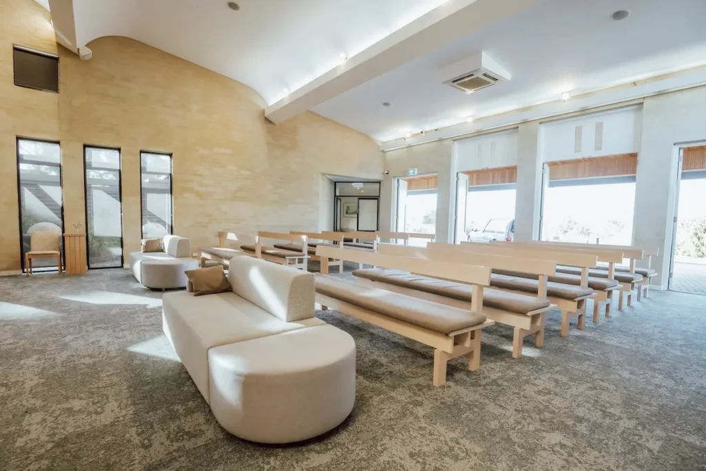 Wide interior view of cushioned timber pews and bright side exit doors inside Geelong Memorial Park Chapel at 42/110 Burvilles Rd, Mount Duneed.