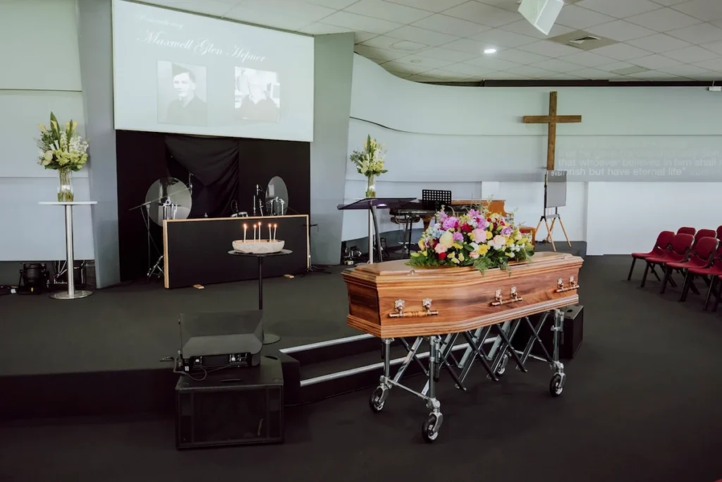Main presentation stage featuring a timber casket with fresh flowers, speaker lectern, candles, and digital photo slideshow screen inside Waurn Ponds Community Chapel in Belmont.